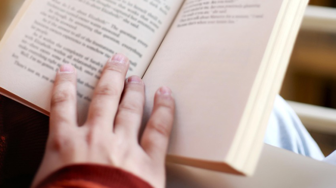 A close-up of a person's hand holding open a book, with the focus on their fingers and the pages. The person is wearing a long-sleeved, reddish-orange top.