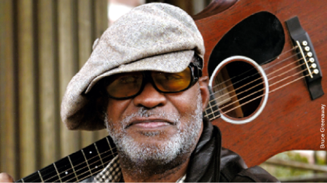 A closeup photograph of an African American man wearing sunglasses and a light gray hat with a guitar over his shoulder. 